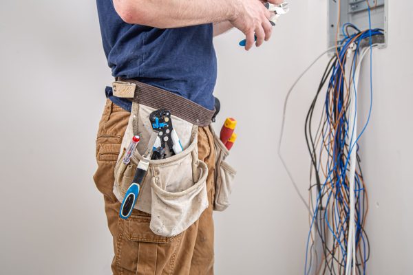 Electrician builder at work, examines the cable connection in the electrical line in the fuselage of an industrial switchboard. Professional in overalls with an electrician's tool.